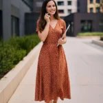 Frau in einem Casual-Outfit für ein Vorstellungsgespräch in einem kreativen oder sozialen Berufsfeld, bestehend aus einem braunen, knielangen Wickelkleid mit weißen Punkten und weißen Sneakern. Sie steht vor modernen Bürogebäuden, lächelt und telefoniert, während sie ein Tablet in der Hand hält. Das Outfit wirkt freundlich, gepflegt und natürlich – ideal für Bewerbungsgespräche in offenen, modernen Arbeitsumgebungen.