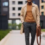 Mann in einem gehobenen Business-Casual-Outfit für ein Vorstellungsgespräch, bestehend aus einem beigen Trenchcoat, einem braunen Rollkragenpullover, grauer Stoffhose und beigen Sneakern. Er trägt eine braune Ledertasche und eine Brille – ein moderner, stilvoller Look, der Professionalität und modisches Gespür ausstrahlt.
