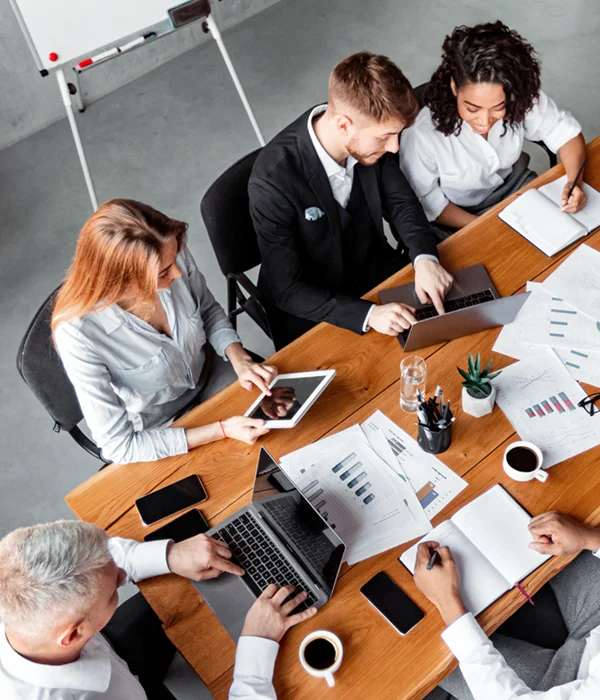 Teammeeting in einer städtischen Organisation: Mehrere Mitarbeitende sitzen an einem großen Tisch und arbeiten gemeinsam an Laptops, Tablets und Unterlagen.