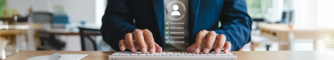 Person in Businesskleidung tippt auf einer Computertastatur, auf der ein digitales Symbol eines Nutzerprofils mit Häkchen eingeblendet ist. Im Hintergrund ist ein helles Büro zu sehen.