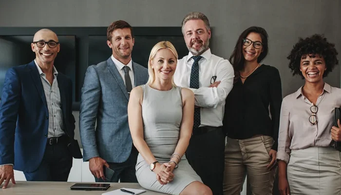 Gruppenfoto eines professionellen Führungsteams in einem modernen Büro: fünf Frauen und Männer in Business-Kleidung stehen selbstbewusst an einem Tisch und lächeln in die Kamera. Das Bild vermittelt Kompetenz, Vielfalt und Führungsstärke – passend zum Thema Executive Search und professionelle Führungskräftevermittlung.