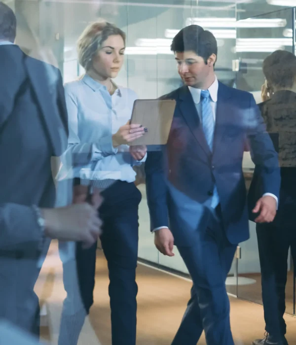 Zwei Geschäftspersonen in formeller Kleidung gehen durch ein modernes Büro und besprechen Dokumente, die auf einem Tablet angezeigt werden. Die Szene symbolisiert professionelle Dienstleistungen im Bereich Steuerberatung und Finanzwesen.