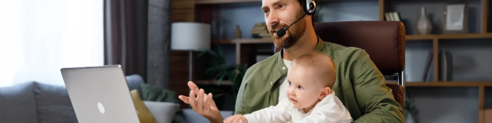 Ein Mann arbeitet im Homeoffice an einem Laptop und hält gleichzeitig ein Baby auf dem Arm. Er trägt ein Headset und führt ein Videomeeting, während er sich um sein Kind kümmert. Das Bild zeigt flexibles Arbeiten und verdeutlicht, wie moderne Arbeitsmodelle die Vereinbarkeit von Beruf und Familie ermöglichen.