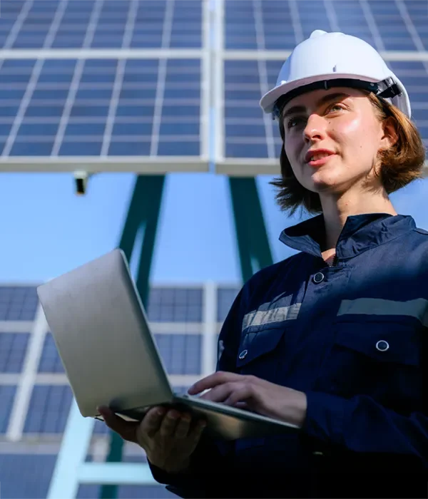 Das Bild zeigt eine Mitarbeiterin von Stadtwerken oder einem Energieversorger, die mit einem Laptop vor einer Solaranlage steht. Sie trägt Schutzkleidung und einen Helm, während sie Daten analysiert oder den Betrieb der Anlage überwacht, was auf ihre Verantwortung in der Wartung und Optimierung von erneuerbaren Energien hinweist.