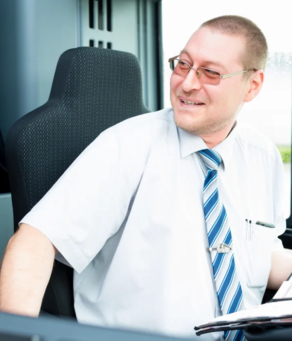 Mitarbeiter im öffentlichen Nahverkehr sitzt am Steuerplatz und lächelt zur Seite. Er trägt Hemd und Krawatte, im Vordergrund liegen Unterlagen. Arbeitssituation aus dem Umfeld von Jobs in öffentlichen Verkehrsbetrieben