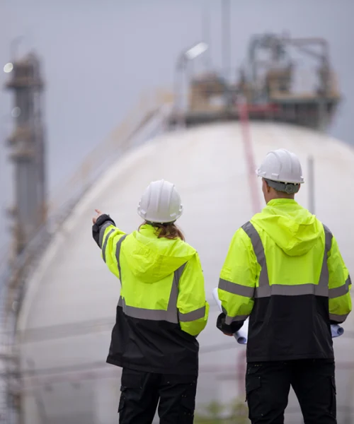 Zwei Personen in Warnschutzkleidung und Helmen stehen vor einer großen Industrieanlage und betrachten die Umgebung, aufgenommen im Arbeitsumfeld der Chemieindustrie.
