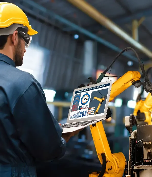 Ein Techniker mit gelbem Schutzhelm und Schutzbrille bedient einen Laptop, der eine Schnittstelle zur Steuerung eines Automationsroboters anzeigt, in einer industriellen Umgebung.