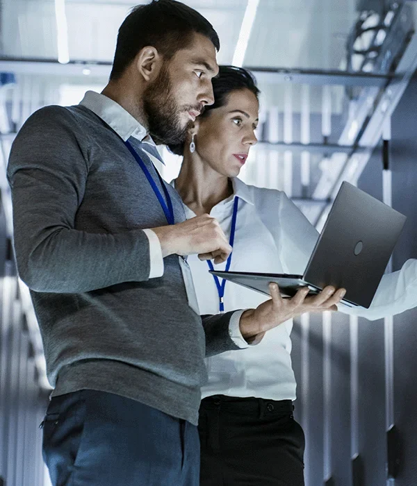 Männlicher IT-Spezialist hält Laptop und bespricht Arbeit mit weiblichem Servertechniker. Sie stehen im Rechenzentrum, der Serverschrank ist geöffnet.