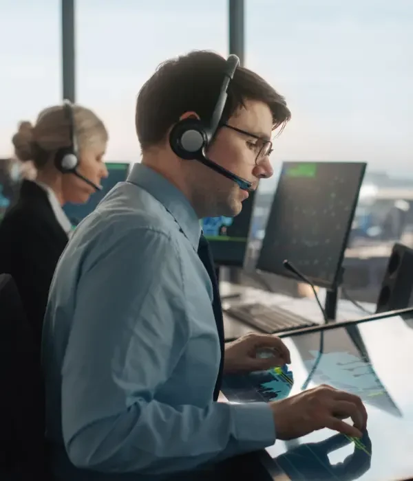 Männlicher Fluglotse mit Headset bei einem Gespräch im Flughafentower. Der Büroraum ist voll mit Desktop-Computern mit Navigationsbildschirmen, Flugzeugabflug- und Ankunftsdaten für das Team.